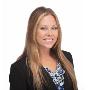 A person with long, light brown hair is smiling and wearing a black blazer over a patterned blouse.