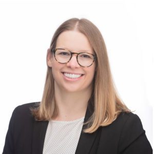 A woman with straight blond hair and glasses is smiling while wearing a black blazer and white top against a plain white background.