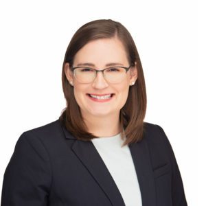 A person with shoulder-length brown hair, glasses, and wearing a dark blazer smiles against a white background.