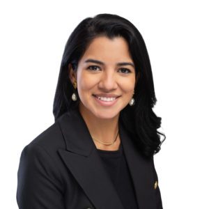 A person with long dark hair is smiling, dressed in a black blazer and a black top, set against a white background.