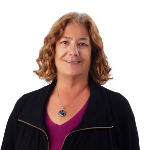 A person with shoulder-length curly hair, wearing a black jacket over a purple top, is smiling at the camera.