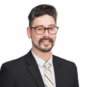A man with short dark hair, a beard, and glasses is wearing a black suit with a white shirt and a striped tie.