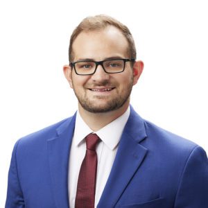 A man wearing glasses, a blue suit, and a burgundy tie is smiling against a white background.