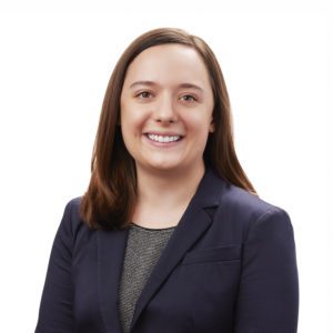 A person with shoulder-length hair is smiling and wearing a dark blazer over a patterned top against a white background.