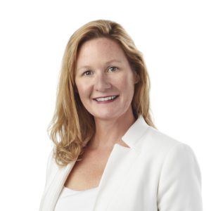 A person with shoulder-length blonde hair is smiling and wearing a white blazer over a white top.