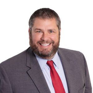 A bearded man is smiling while wearing a grey suit with a red tie.