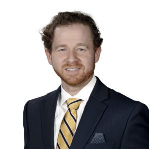 A person with curly hair wearing a dark suit, white shirt, and striped yellow tie smiles against a plain white background.
