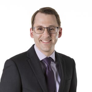 A person wearing glasses and dressed in a suit with a tie is smiling against a plain white background.