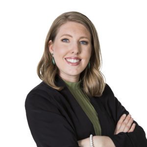 A person with shoulder-length hair is smiling, wearing a black blazer over a green top, and has their arms crossed.