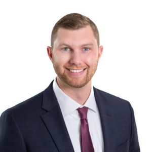 A man in a navy suit and maroon tie is smiling against a white background.