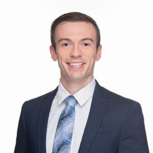 A person is wearing a suit, white shirt, and patterned tie, smiling against a plain white background.