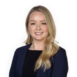 A person with long blonde hair wearing a dark blazer and a black top smiles against a white background.