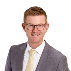 A person with glasses and red hair is wearing a gray suit and a yellow tie, smiling against a white background.