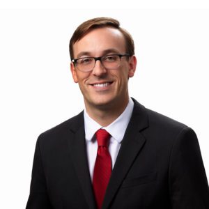 A person with glasses is wearing a suit and red tie and smiling against a white background.