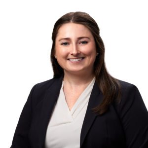 A person with long brown hair is smiling and wearing a black blazer over a white top.