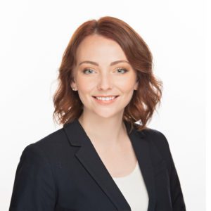 A person with short auburn hair is wearing a dark blazer and smiling against a plain white background.