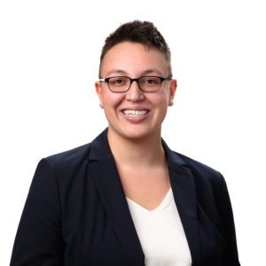 A person with short hair, wearing glasses, a navy blazer, and a white shirt, is smiling against a white background.