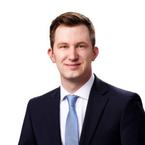 A person dressed in a suit and tie is smiling against a plain white background.