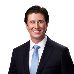 A person wearing a dark suit, white shirt, and light blue tie is smiling against a plain white background.