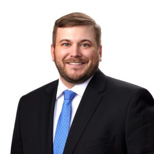 A man in a black suit and blue tie smiling against a white background.
