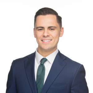 A person in a suit and tie is smiling against a plain white background.