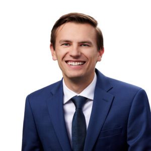 A man in a blue suit and tie is smiling against a white background.