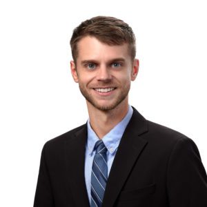 A man with short brown hair and a trimmed beard is wearing a suit, light blue shirt, and striped tie, smiling against a white background.