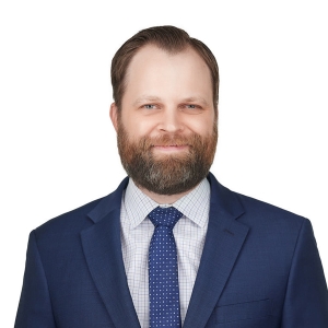 A bearded man in a blue suit and checkered tie poses against a plain white background.