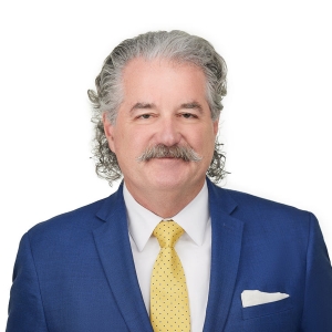 A man with gray, curly hair and a mustache is wearing a blue suit, a white shirt, a yellow tie with polka dots, and a white pocket square.