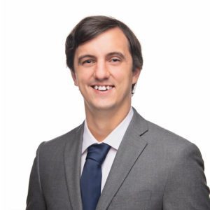 A smiling individual is dressed in a grey suit with a blue tie against a plain white background.