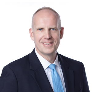 A person is wearing a dark suit, light blue shirt, and pink tie, posing against a plain white background.