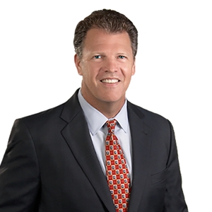 A man wearing a dark suit, light blue shirt, and a patterned red tie smiles against a white background.