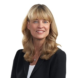 A woman with long blonde hair and bangs is smiling while wearing a black blazer over a white top.