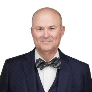A bald man wearing a dark suit, white dress shirt, and a striped bow tie is smiling against a white background.