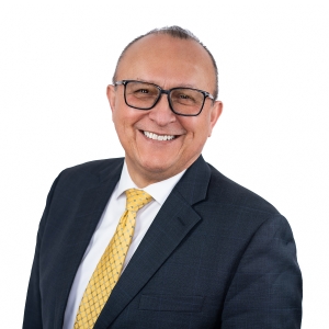 A person wearing a dark suit, white shirt, and yellow tie, smiling against a plain white background.