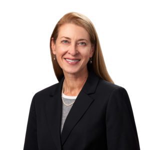 A person with long blond hair is wearing a dark blazer over a light shirt, smiling at the camera against a plain white background.