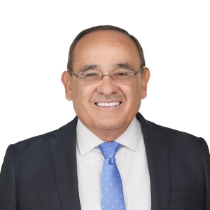 A man with glasses is smiling, wearing a dark suit, white shirt, and a blue tie.