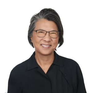 A person with short, black hair speckled with gray is smiling, wearing glasses and a black collared shirt against a white background.