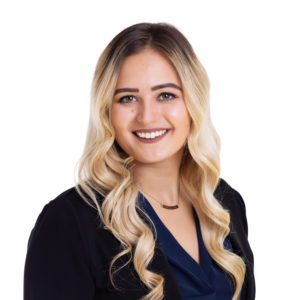 A woman with long, wavy blonde hair is smiling and wearing a dark blazer over a blue top.