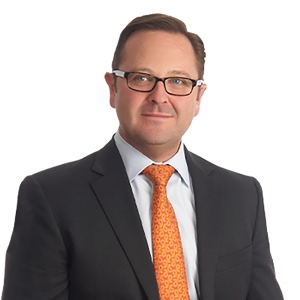 A man wearing glasses, a suit, and an orange tie smiles against a white background.
