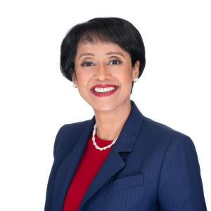 A woman wearing a blue suit jacket, red top, and pearl necklace is smiling against a white background.