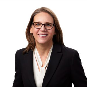 A person with glasses is wearing a dark blazer and a white blouse, smiling against a pure white background.