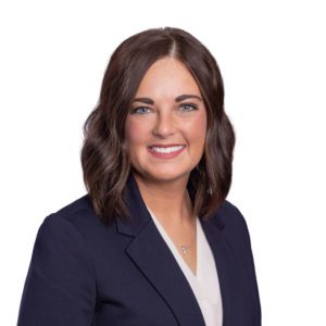 A person with shoulder-length brown hair is smiling, wearing a navy blazer and white top against a white background.