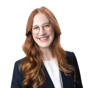 A smiling individual with long, wavy, red hair wearing glasses and a dark blazer over a white top.