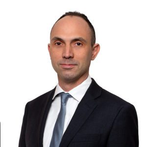 A man in a black suit and light blue tie stands against a plain white background.