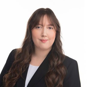 A person with long brown hair and bangs is wearing a black blazer and white top, smiling slightly against a white background.