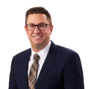 A man wearing glasses, a navy suit, a white shirt, and a striped tie is smiling against a plain white background.
