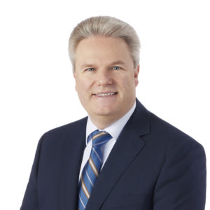 A man in a suit and blue striped tie is smiling against a plain white background.
