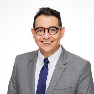 A person with short dark hair and glasses is smiling, wearing a gray suit, white shirt, and a blue tie against a white background.