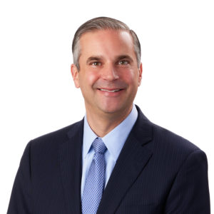 A man in a suit and tie smiles against a plain white background.